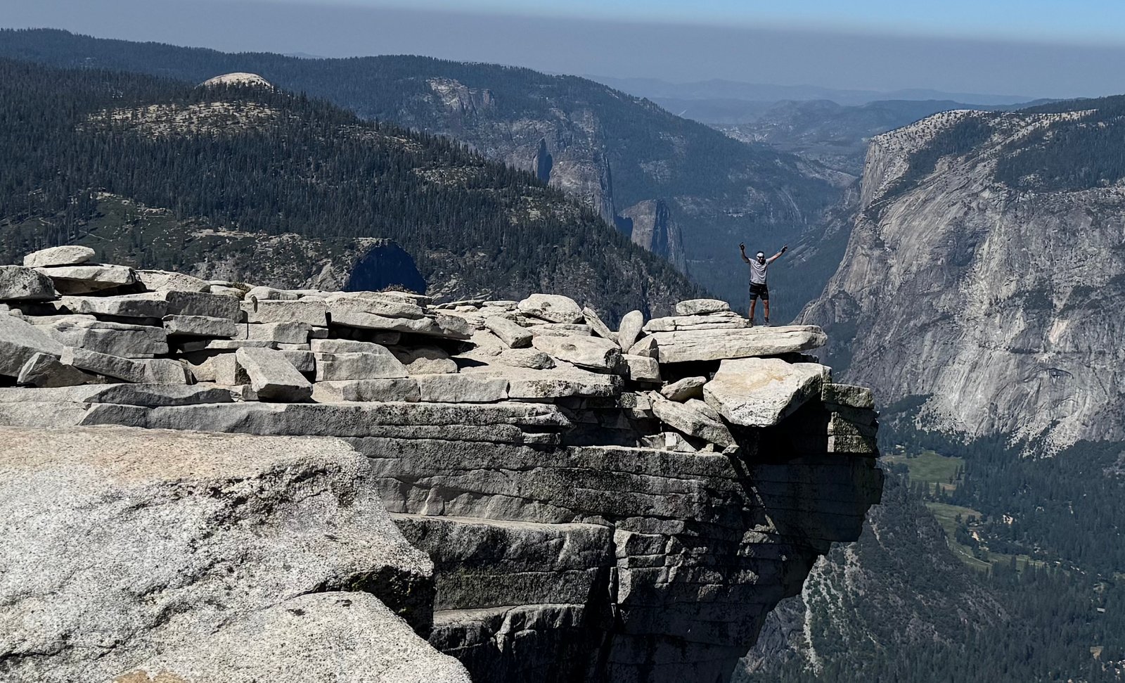 Daniel Greenwood standing on the ledge at the summit, arms raised, with the valley behind him